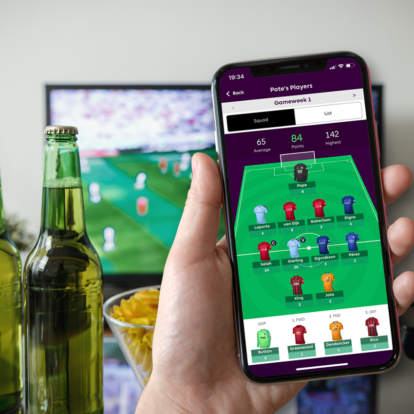 Someone holding a phone displaying Fantasy Premier League app on it, whilst football plays on a TV in the background